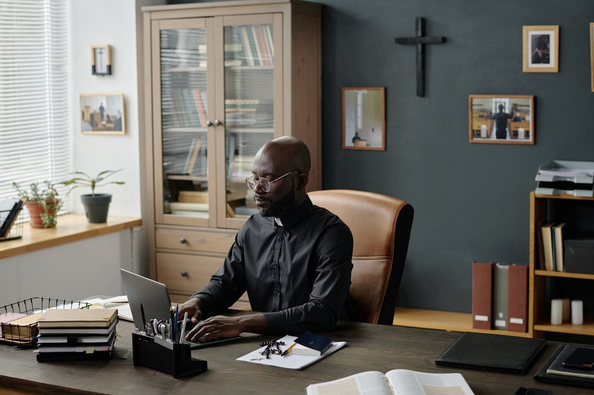 Wide shot of spacious office with African American pastor working on laptop sitting at wooden table