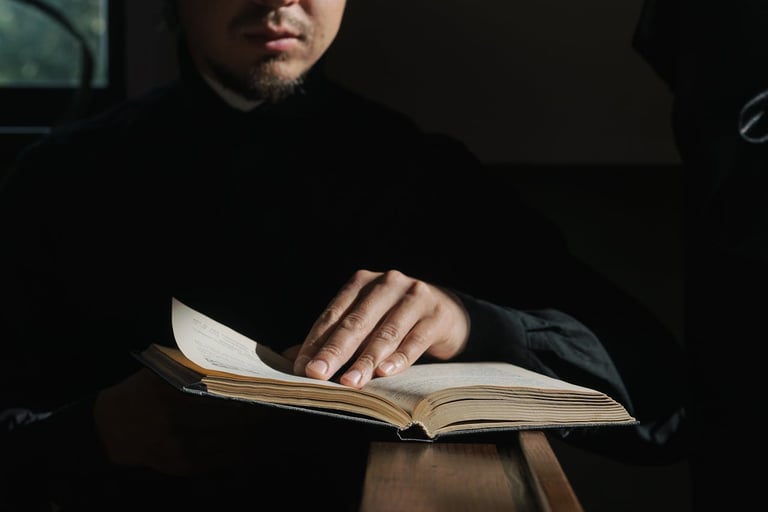 Man in dark clothing reading an open book against a dark background, with his hand turning the page